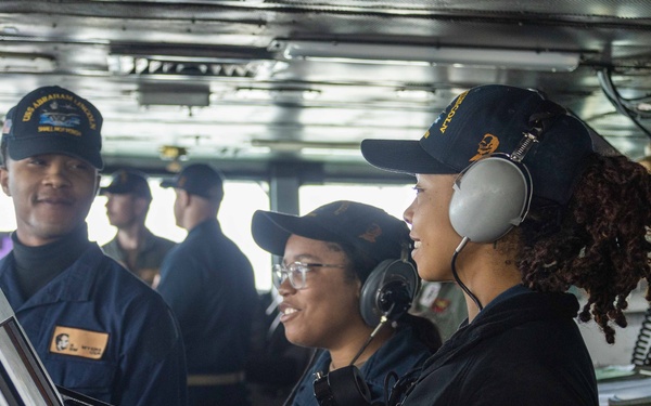 The image shows U.S. Navy personnel on the bridge of what appears to be USS Abraham Lincoln, a naval vessel. Three sailors are prominently featured in the foreground, with one wearing communication headsets and another wearing glasses, all dressed in Navy working uniforms with ship-specific caps. Additional crew members are visible in the background, suggesting an active watch or operational period.
