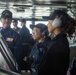 The image shows U.S. Navy personnel on the bridge of what appears to be USS Abraham Lincoln, a naval vessel. Three sailors are prominently featured in the foreground, with one wearing communication headsets and another wearing glasses, all dressed in Navy working uniforms with ship-specific caps. Additional crew members are visible in the background, suggesting an active watch or operational period.