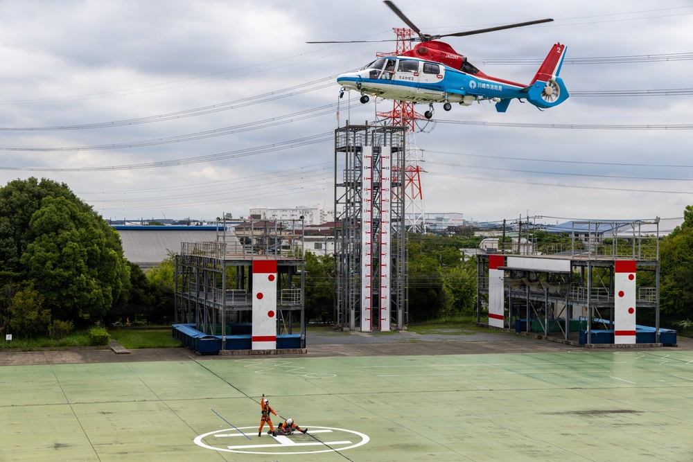 A red, white, and blue helicopter marked with Japanese text (川崎市消防局 - Kawasaki City Fire Department) hovers over a training facility while conducting a rescue hoist exercise. A rescue worker in orange gear on the ground appears to be connected to a cable lowered from the helicopter, with what appears to be a simulated casualty nearby. The scene takes place at what is clearly a dedicated emergency services training ground with marked helipads and training structures.