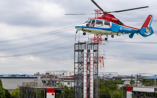 A red, white, and blue helicopter marked with Japanese text (川崎市消防局 - Kawasaki City Fire Department) hovers over a training facility while conducting a rescue hoist exercise. A rescue worker in orange gear on the ground appears to be connected to a cable lowered from the helicopter, with what appears to be a simulated casualty nearby. The scene takes place at what is clearly a dedicated emergency services training ground with marked helipads and training structures.