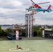 A red, white, and blue helicopter marked with Japanese text (川崎市消防局 - Kawasaki City Fire Department) hovers over a training facility while conducting a rescue hoist exercise. A rescue worker in orange gear on the ground appears to be connected to a cable lowered from the helicopter, with what appears to be a simulated casualty nearby. The scene takes place at what is clearly a dedicated emergency services training ground with marked helipads and training structures.