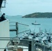 The image shows the flight deck of a US Navy aircraft carrier packed with folded-wing fighter jets, viewed from an elevated catwalk or island structure. A single person in dark clothing leans against a railing observing the harbor scene. A red-and-white tugboat navigates the calm harbor waters, with a green, hilly island and port facilities visible in the background.
