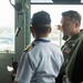 The image shows a group of military personnel in what appears to be a ship's bridge or command center. A US Navy officer in a green flight suit with an American flag patch is conversing with another officer in a white naval uniform with gold shoulder epaulettes. The setting suggests an official military exchange or tour aboard a naval vessel.