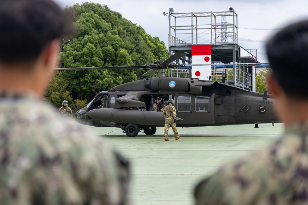 A U.S. Army UH-60 Black Hawk helicopter is parked on a landing pad, with American soldiers in MultiCam uniforms conducting ground operations around it. Japanese military personnel in camouflage uniforms are visible in the foreground observing the aircraft, suggesting a joint military exercise or demonstration. A structure bearing red-and-white markings consistent with Japanese flag colors is visible in the background.