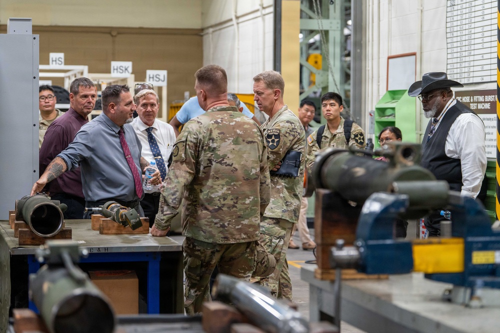 A group of military personnel in Army Combat Uniforms and civilian officials are conducting a facility tour or briefing inside what appears to be a military depot or manufacturing/testing facility. A civilian in a gray shirt and red tie appears to be presenting or explaining equipment on a workbench. Various munitions components and industrial testing machinery are visible throughout the space.