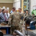 A group of military personnel in Army Combat Uniforms and civilian officials are conducting a facility tour or briefing inside what appears to be a military depot or manufacturing/testing facility. A civilian in a gray shirt and red tie appears to be presenting or explaining equipment on a workbench. Various munitions components and industrial testing machinery are visible throughout the space.
