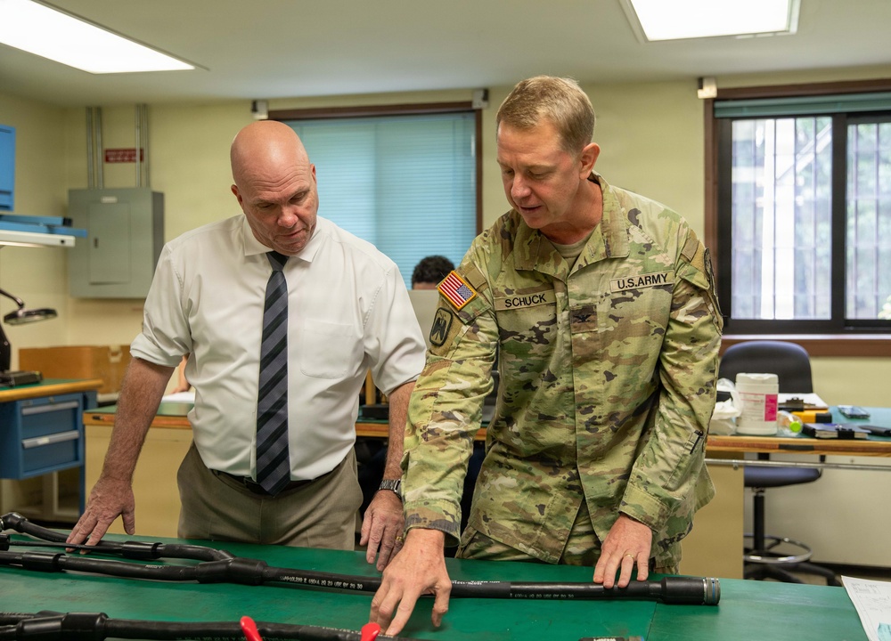 A U.S. Army soldier identified by name tape as 'SCHUCK' is showing or explaining equipment laid out on a green-surfaced workbench to a civilian man in a white dress shirt and striped tie. The setting appears to be a military laboratory or technical workspace. Both individuals are leaning over the table examining what appear to be weapon components or technical equipment.