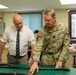 A U.S. Army soldier identified by name tape as 'SCHUCK' is showing or explaining equipment laid out on a green-surfaced workbench to a civilian man in a white dress shirt and striped tie. The setting appears to be a military laboratory or technical workspace. Both individuals are leaning over the table examining what appear to be weapon components or technical equipment.