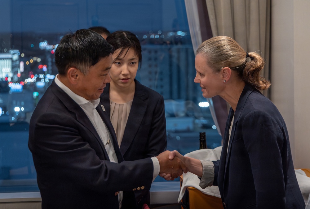 A formal meeting or greeting is taking place indoors, likely in a hotel or conference room setting at night. Two individuals in the foreground are shaking hands — a man in a dark suit on the left and a woman in a dark blazer on the right. A younger woman in a dark blazer stands between them, appearing to observe the exchange, possibly serving as an interpreter or aide.