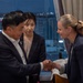 A formal meeting or greeting is taking place indoors, likely in a hotel or conference room setting at night. Two individuals in the foreground are shaking hands — a man in a dark suit on the left and a woman in a dark blazer on the right. A younger woman in a dark blazer stands between them, appearing to observe the exchange, possibly serving as an interpreter or aide.