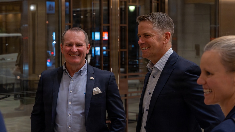 Two men in dark business suits are standing together in what appears to be a modern building lobby or atrium, both smiling and appearing engaged in conversation. A third person, partially visible on the right edge, also appears to be smiling. The setting suggests a professional or formal social gathering.