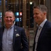 Two men in dark business suits are standing together in what appears to be a modern building lobby or atrium, both smiling and appearing engaged in conversation. A third person, partially visible on the right edge, also appears to be smiling. The setting suggests a professional or formal social gathering.