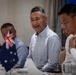 Three men are seated at a dining table in what appears to be a formal meeting or banquet setting. A small Australian flag is visible in the foreground, suggesting a diplomatic, business, or official gathering. The central figure is smiling and appears engaged in conversation.
