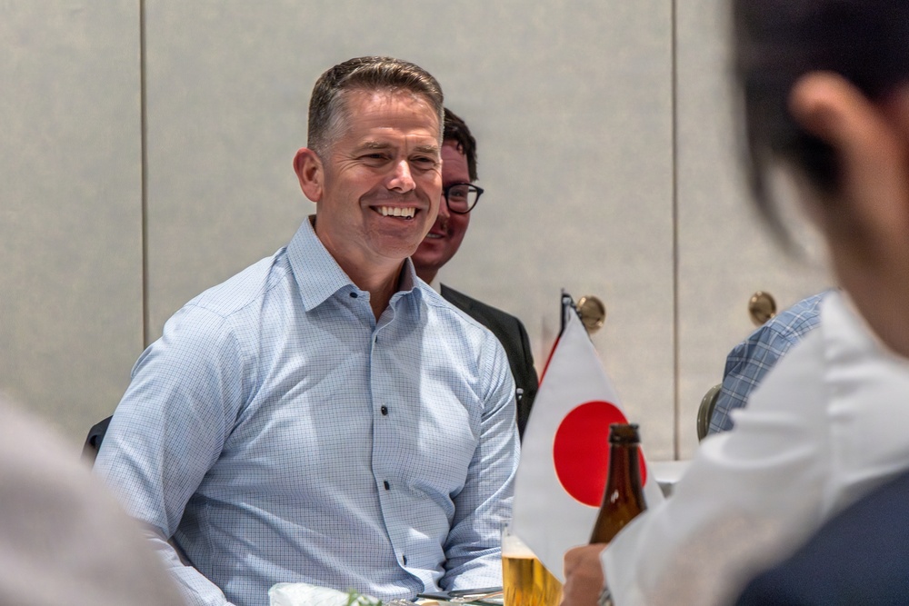 A smiling man in a light blue checkered dress shirt sits at a table during what appears to be a formal or semi-formal meeting or dining event. A small Japanese national flag (Hinomaru) is visible on the table, suggesting a Japan-related official or diplomatic context. Several other individuals are partially visible in the foreground and background.