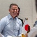 A smiling man in a light blue checkered dress shirt sits at a table during what appears to be a formal or semi-formal meeting or dining event. A small Japanese national flag (Hinomaru) is visible on the table, suggesting a Japan-related official or diplomatic context. Several other individuals are partially visible in the foreground and background.