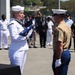 A formal military ceremony is taking place outdoors, likely a flag-folding or memorial service. A U.S. Navy sailor in dress whites is presenting a folded American flag to a U.S. Marine in service uniform (tan blouse, blue trousers with red stripe). A multinational audience of military personnel and civilians observes in the background.