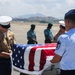 A solemn military dignified transfer ceremony is taking place on an airfield tarmac. A U.S. Marine Corps Staff Sergeant and a U.S. Air Force Master Sergeant are carefully draping or folding an American flag over a flag-draped transfer case (casket). Foreign military personnel in teal/green uniforms stand at attention in the background as witnesses.