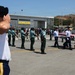 A formal military repatriation ceremony is taking place on an airport tarmac, with a flag-draped casket being carried by uniformed pallbearers. The setting appears to be Port Moresby's Jackson's International Airport in Papua New Guinea, based on the visible signage. Personnel from multiple military branches and nations are present, rendering salutes as the casket passes.