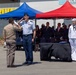 A formal military ceremony is taking place outdoors at what appears to be an airport arrivals area, likely involving the repatriation of remains. Two military personnel are exchanging salutes — one in U.S. Air Force dress blues and one in a tan/khaki uniform — while a U.S. Navy sailor in dress whites stands at attention beside a flag-draped or covered object on a table. A crowd of military and civilian dignitaries observes in the background.