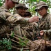 A group of U.S. Army soldiers in MultiCam camouflage uniforms are engaged in a field training exercise involving rope work or knot-tying around a log or tree stump in a wooded outdoor environment. At least one soldier's name tape reads 'DELGADO,' and another's uniform displays a unit patch consistent with the 2nd Infantry Division. The soldiers appear focused on a hands-on task, suggesting a survival, engineering, or rappelling skills training scenario.