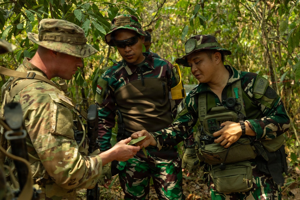 Three military personnel are gathered in a forested environment, appearing to examine a small object or document. One soldier wearing US Army MultiCam (OCP) uniform is interacting with two soldiers wearing Indonesian Armed Forces (TNI) woodland camouflage uniforms, suggesting a joint military training or coordination exercise. The setting and interaction indicate a bilateral military cooperation activity in a jungle or tropical environment.