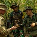 Three military personnel are gathered in a forested environment, appearing to examine a small object or document. One soldier wearing US Army MultiCam (OCP) uniform is interacting with two soldiers wearing Indonesian Armed Forces (TNI) woodland camouflage uniforms, suggesting a joint military training or coordination exercise. The setting and interaction indicate a bilateral military cooperation activity in a jungle or tropical environment.