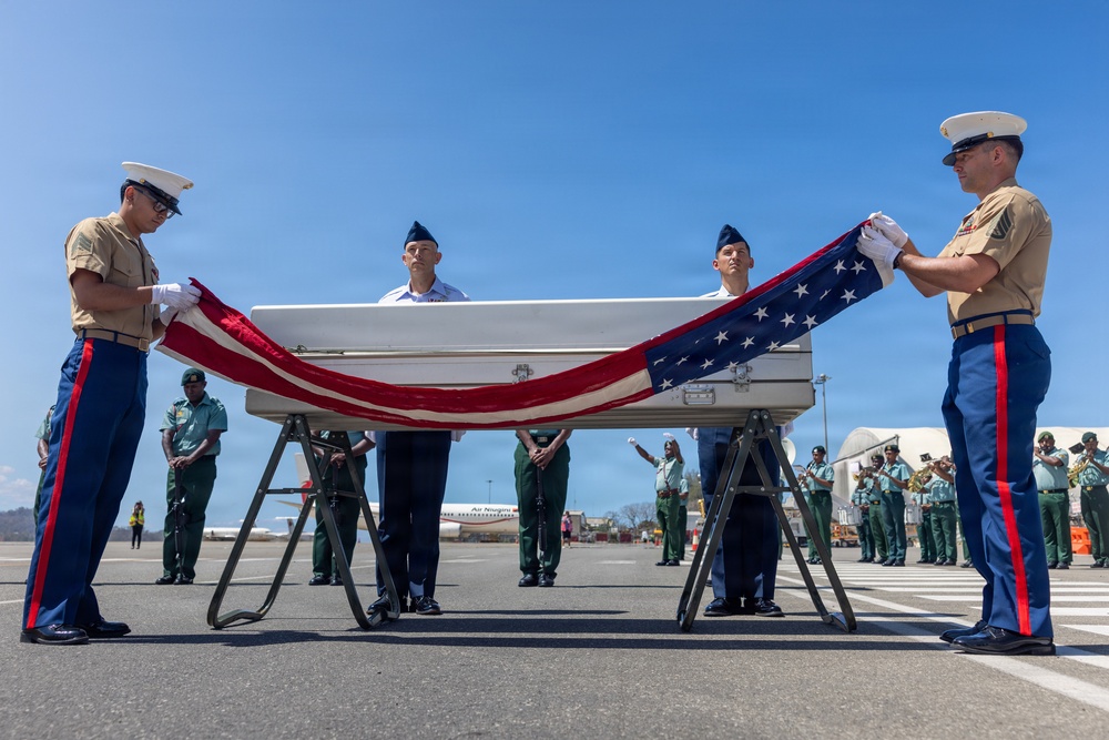 A military honors ceremony is taking place on an airport tarmac, where U.S. Marines and Air Force personnel are draping an American flag over a white transfer case (casket). The ceremony appears to be a dignified transfer of remains, conducted with full military honors. Foreign military personnel and a band are visible in formation in the background, suggesting an international repatriation ceremony.
