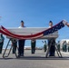 A military honors ceremony is taking place on an airport tarmac, where U.S. Marines and Air Force personnel are draping an American flag over a white transfer case (casket). The ceremony appears to be a dignified transfer of remains, conducted with full military honors. Foreign military personnel and a band are visible in formation in the background, suggesting an international repatriation ceremony.