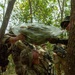 A U.S. Army soldier in MultiCam/OCP uniform is drinking liquid from a large plastic bag filled with green vegetation, likely a jungle survival training exercise. The bag appears to contain plant material from which water or liquid is being extracted, consistent with wilderness survival water-collection techniques. Other soldiers and what appears to be a foreign military instructor in a jungle hat are assisting and observing.