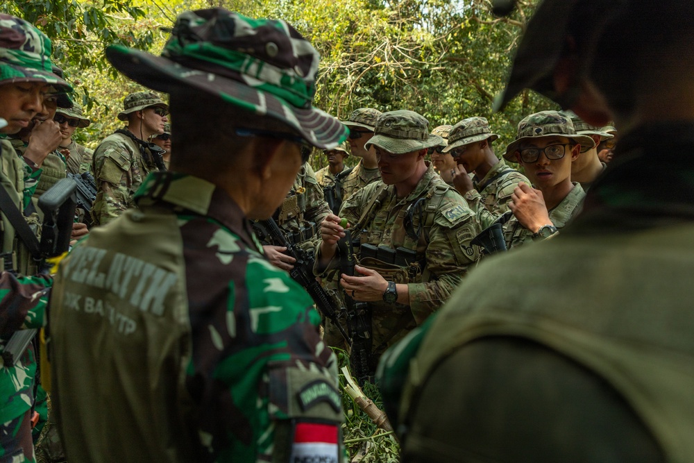 A group of military personnel from at least two different nations are gathered in a jungle or forested environment, appearing to be engaged in a joint training exercise. Soldiers wearing Indonesian TNI (Tentara Nasional Indonesia) camouflage uniforms are interacting with soldiers in MultiCam/OCP uniforms, likely US Army personnel. One soldier is handling a rifle, possibly demonstrating a technique or receiving instruction.