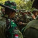 A group of military personnel from at least two different nations are gathered in a jungle or forested environment, appearing to be engaged in a joint training exercise. Soldiers wearing Indonesian TNI (Tentara Nasional Indonesia) camouflage uniforms are interacting with soldiers in MultiCam/OCP uniforms, likely US Army personnel. One soldier is handling a rifle, possibly demonstrating a technique or receiving instruction.