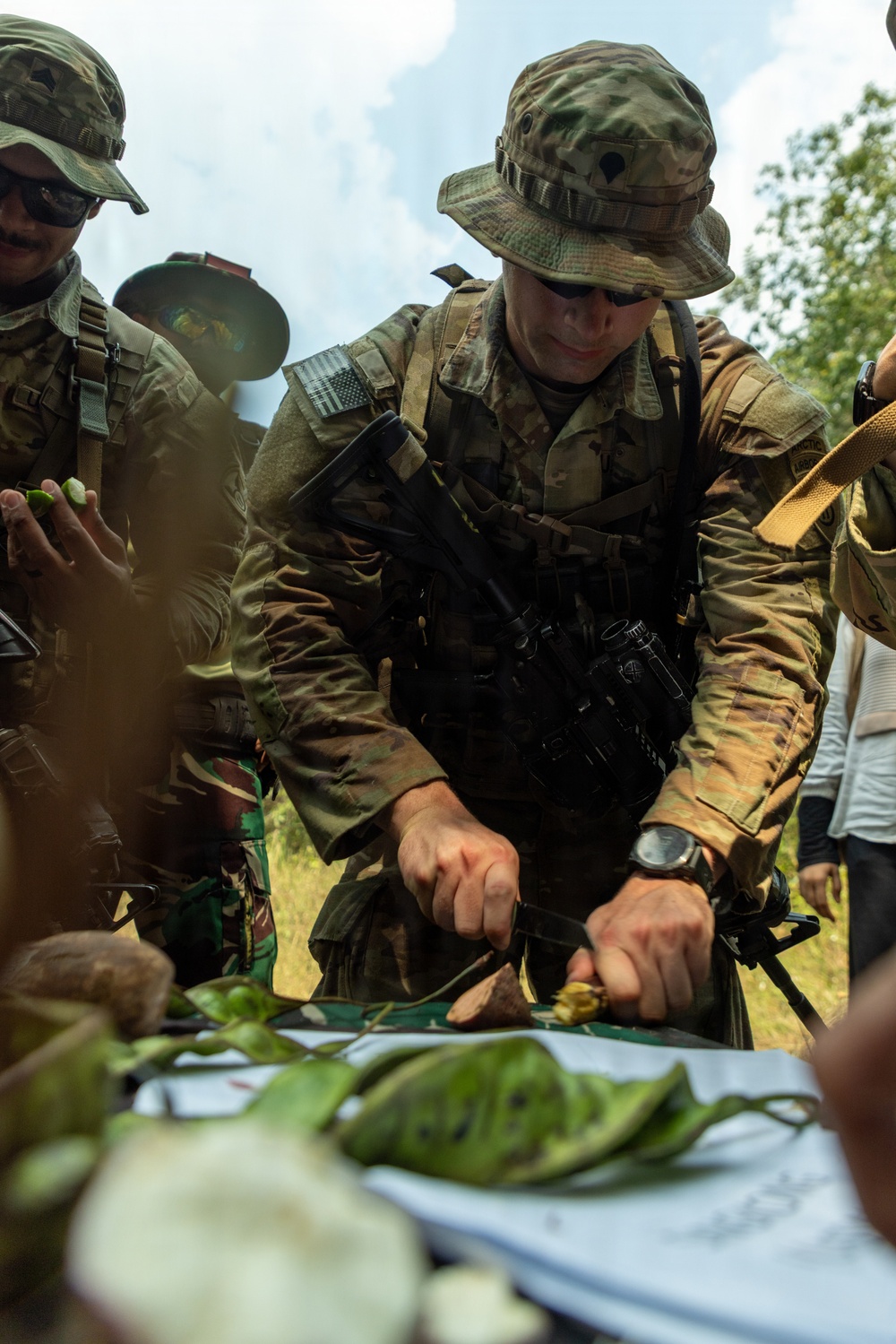 A U.S. Army soldier in MultiCam/OCP uniform and boonie hat is using a knife to cut or prepare vegetation/food items on a surface covered with large leaves. He is accompanied by at least one other soldier wearing a different camouflage pattern, suggesting a joint or multinational training exercise. The setting appears to be an outdoor, tropical or jungle environment.