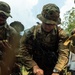 A U.S. Army soldier in MultiCam/OCP uniform and boonie hat is using a knife to cut or prepare vegetation/food items on a surface covered with large leaves. He is accompanied by at least one other soldier wearing a different camouflage pattern, suggesting a joint or multinational training exercise. The setting appears to be an outdoor, tropical or jungle environment.