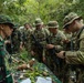 A group of military personnel in camouflage uniforms are gathered around a table covered with various jungle plants and root vegetables during what appears to be a jungle survival or field craft training exercise. Two distinct uniform patterns are visible, suggesting a joint multinational military exercise, with one group wearing Indonesian TNI (Tentara Nasional Indonesia) camouflage and the other wearing US Army Operational Camouflage Pattern (OCP). The soldiers are examining and handling edible plants laid out on a camouflage-covered table with identification labels.
