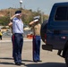 Three military personnel in dress uniforms are rendering a formal salute toward a dark blue SUV/vehicle on what appears to be a tarmac or paved area. Two individuals wear U.S. Air Force-style light blue shirts with dark navy trousers featuring red stripes, while the third wears U.S. Marine Corps tan service uniform with a white cover. The setting appears to be an outdoor military ceremony, possibly an arrival or departure honors event.