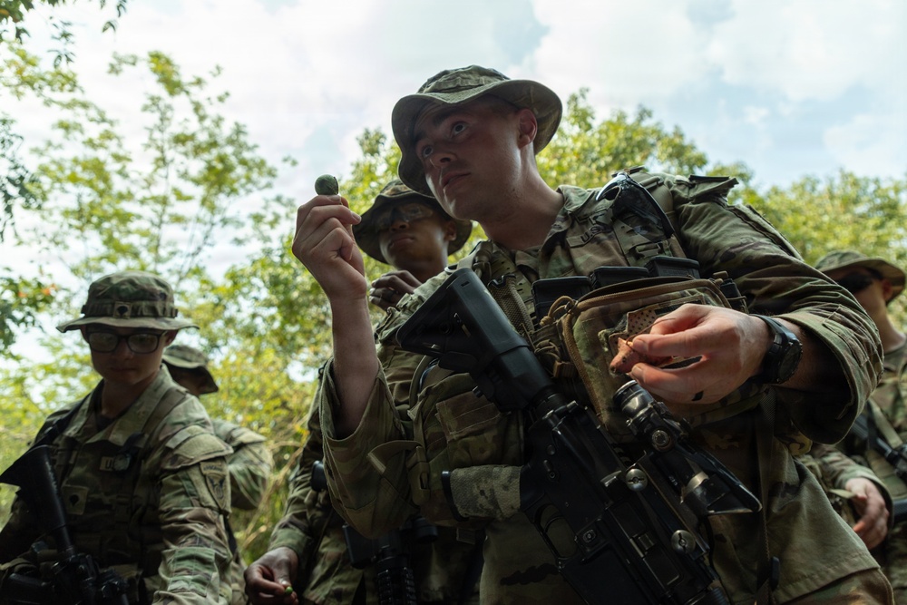 A group of military personnel in full combat gear and camouflage uniforms are gathered in a wooded outdoor setting. The foreground soldier is holding up a small dark object for close inspection, possibly during a field training exercise involving survival or land navigation skills. Multiple soldiers carrying rifles and tactical vests are visible in the background.