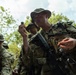 A group of military personnel in full combat gear and camouflage uniforms are gathered in a wooded outdoor setting. The foreground soldier is holding up a small dark object for close inspection, possibly during a field training exercise involving survival or land navigation skills. Multiple soldiers carrying rifles and tactical vests are visible in the background.