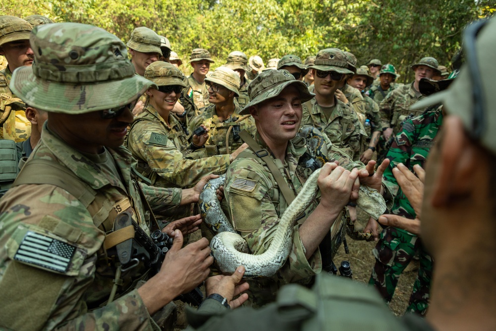A group of military personnel in camouflage uniforms are gathered outdoors in a wooded area, handling a large snake, likely during a jungle survival training exercise. The group includes soldiers wearing US Army multicam uniforms with American flag patches, as well as at least one soldier in Indonesian-pattern camouflage, suggesting a joint multinational military exercise. The central soldier is holding the snake while others crowd around with expressions of curiosity and amusement.