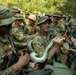 A group of military personnel in camouflage uniforms are gathered outdoors in a wooded area, handling a large snake, likely during a jungle survival training exercise. The group includes soldiers wearing US Army multicam uniforms with American flag patches, as well as at least one soldier in Indonesian-pattern camouflage, suggesting a joint multinational military exercise. The central soldier is holding the snake while others crowd around with expressions of curiosity and amusement.