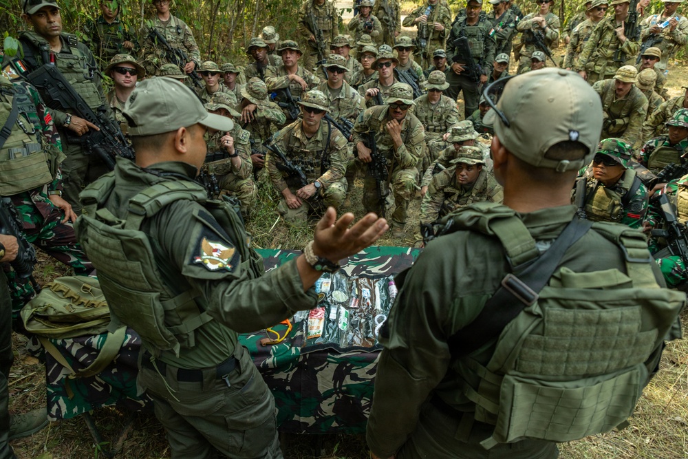 A multinational military training exercise or briefing is taking place in an outdoor jungle/forest setting. Two instructors in olive-green uniforms and caps are conducting a demonstration or briefing in front of a large group of soldiers wearing mixed camouflage uniforms. Items are laid out on a camouflage cloth on the ground, suggesting a field equipment or medical demonstration.