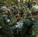 A multinational military training exercise or briefing is taking place in an outdoor jungle/forest setting. Two instructors in olive-green uniforms and caps are conducting a demonstration or briefing in front of a large group of soldiers wearing mixed camouflage uniforms. Items are laid out on a camouflage cloth on the ground, suggesting a field equipment or medical demonstration.