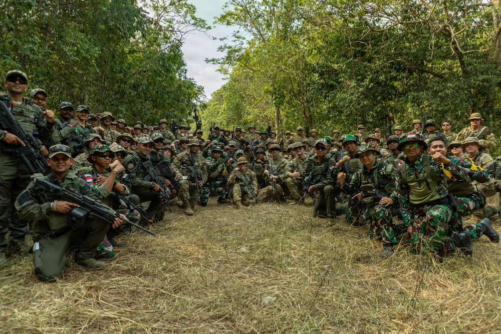 A large group of multinational military personnel pose together for a group photograph in a forested outdoor training area. The soldiers are wearing a mix of camouflage uniforms and carrying various weapons, suggesting a joint military exercise or training event. The presence of Indonesian flag patches and Western-style multicam uniforms indicates a multinational military cooperation exercise.