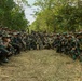 A large group of multinational military personnel pose together for a group photograph in a forested outdoor training area. The soldiers are wearing a mix of camouflage uniforms and carrying various weapons, suggesting a joint military exercise or training event. The presence of Indonesian flag patches and Western-style multicam uniforms indicates a multinational military cooperation exercise.
