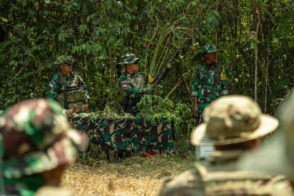 The image shows military personnel in a jungle or forested outdoor setting, appearing to conduct a training or instructional session. The central figure is demonstrating camouflage techniques using natural vegetation, holding leafy branches over a table covered with camouflage netting. Multiple soldiers wearing Indonesian Armed Forces (TNI) uniforms are present, with observers in the foreground watching the demonstration.