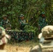The image shows military personnel in a jungle or forested outdoor setting, appearing to conduct a training or instructional session. The central figure is demonstrating camouflage techniques using natural vegetation, holding leafy branches over a table covered with camouflage netting. Multiple soldiers wearing Indonesian Armed Forces (TNI) uniforms are present, with observers in the foreground watching the demonstration.
