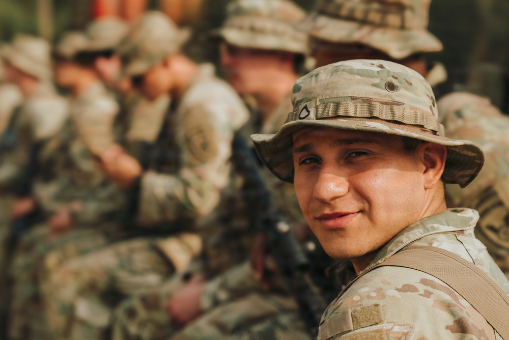 A young soldier in the foreground is looking back over his shoulder toward the camera with a slight smile. He is wearing MultiCam (OCP) pattern camouflage uniform and a boonie hat. Behind him, several other soldiers in similar uniforms are visible but out of focus.