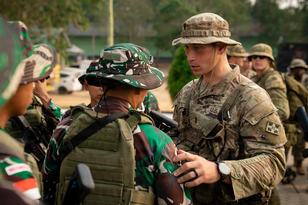 The image shows a multinational military exercise or joint training event involving U.S. Army soldiers and what appear to be Indonesian Armed Forces (TNI) personnel, identifiable by their distinctive green camouflage uniforms and boonie hats. A U.S. soldier wearing a '4th Infantry Division' patch and 'U.S.' chest tape is interacting with foreign military counterparts, possibly reviewing equipment or conducting a briefing. Additional personnel in Australian-style camouflage are visible in the background.