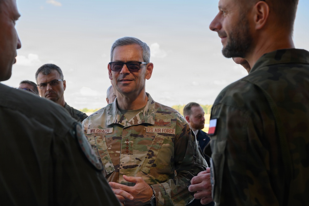 A U.S. Air Force officer identified by name tape as 'WILSBACH' with four-star rank insignia is engaged in conversation with other military personnel. The central figure wears OCP (Operational Camouflage Pattern) uniform with 'U.S. AIR FORCE' tape and sunglasses. At least one individual in the background wears a uniform with what appears to be a Polish flag patch, suggesting a multinational military engagement.
