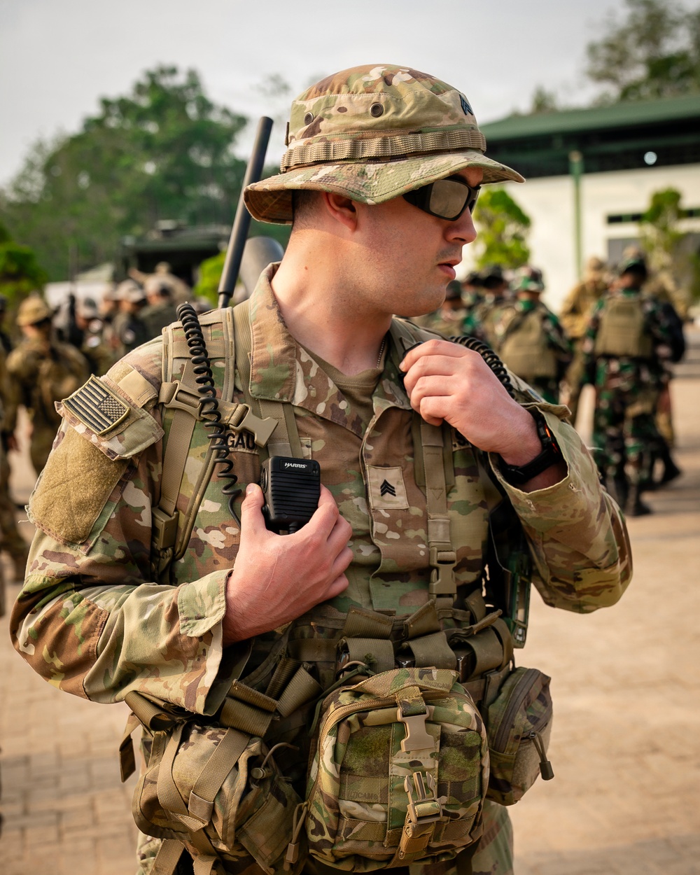 A U.S. Army Sergeant in full MultiCam (OCP) combat gear is holding a Harris radio handset, appearing to communicate during what looks like a joint military exercise or operation. He is wearing a boonie hat, tactical sunglasses, body armor, and a full kit with pouches and a backpack-mounted radio antenna. In the background, soldiers in different uniforms — including what appear to be Indonesian Army (TNI-AD) green camouflage uniforms — are visible, suggesting a multinational training exercise.