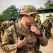 A U.S. Army Sergeant in full MultiCam (OCP) combat gear is holding a Harris radio handset, appearing to communicate during what looks like a joint military exercise or operation. He is wearing a boonie hat, tactical sunglasses, body armor, and a full kit with pouches and a backpack-mounted radio antenna. In the background, soldiers in different uniforms — including what appear to be Indonesian Army (TNI-AD) green camouflage uniforms — are visible, suggesting a multinational training exercise.